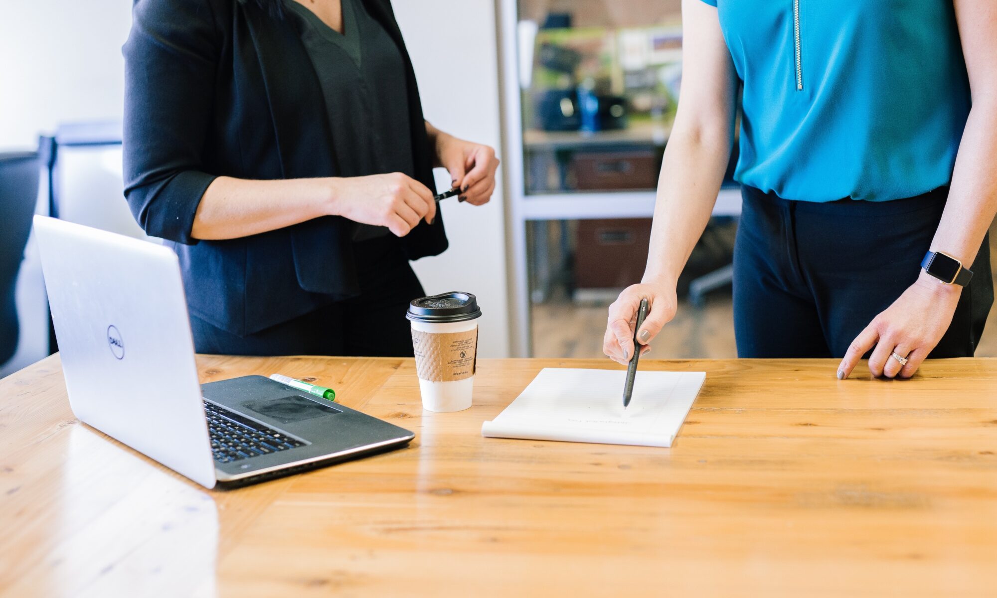 Two women stand in front of a table with a laptop and a notepad