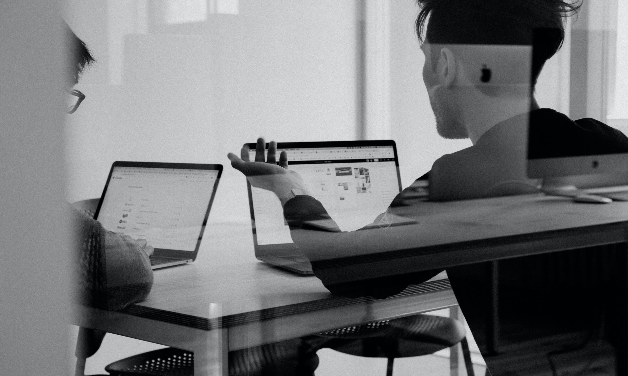 Two men sit in front of laptops discussing something in black and white.