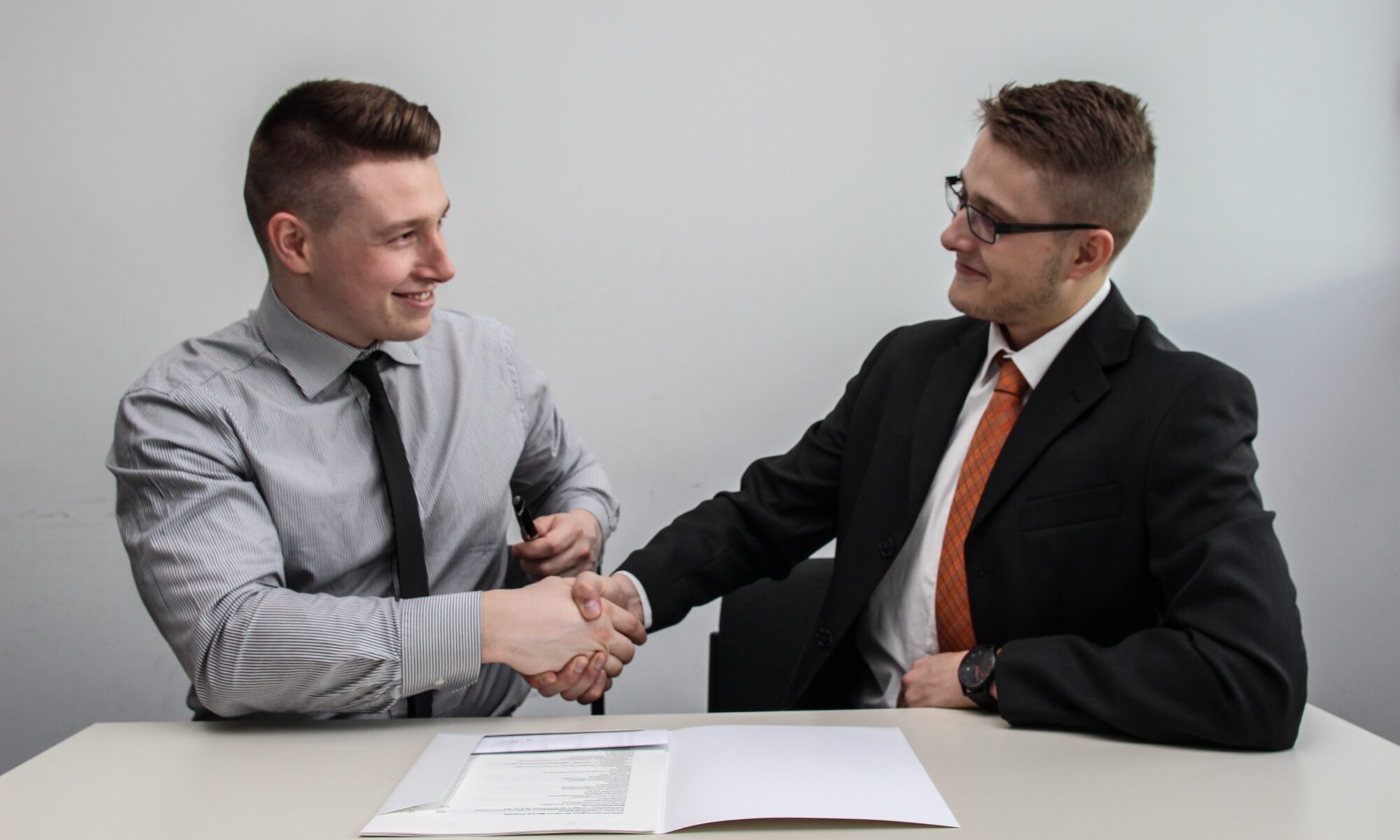 Two men in suits shake hands in front of papers on a table