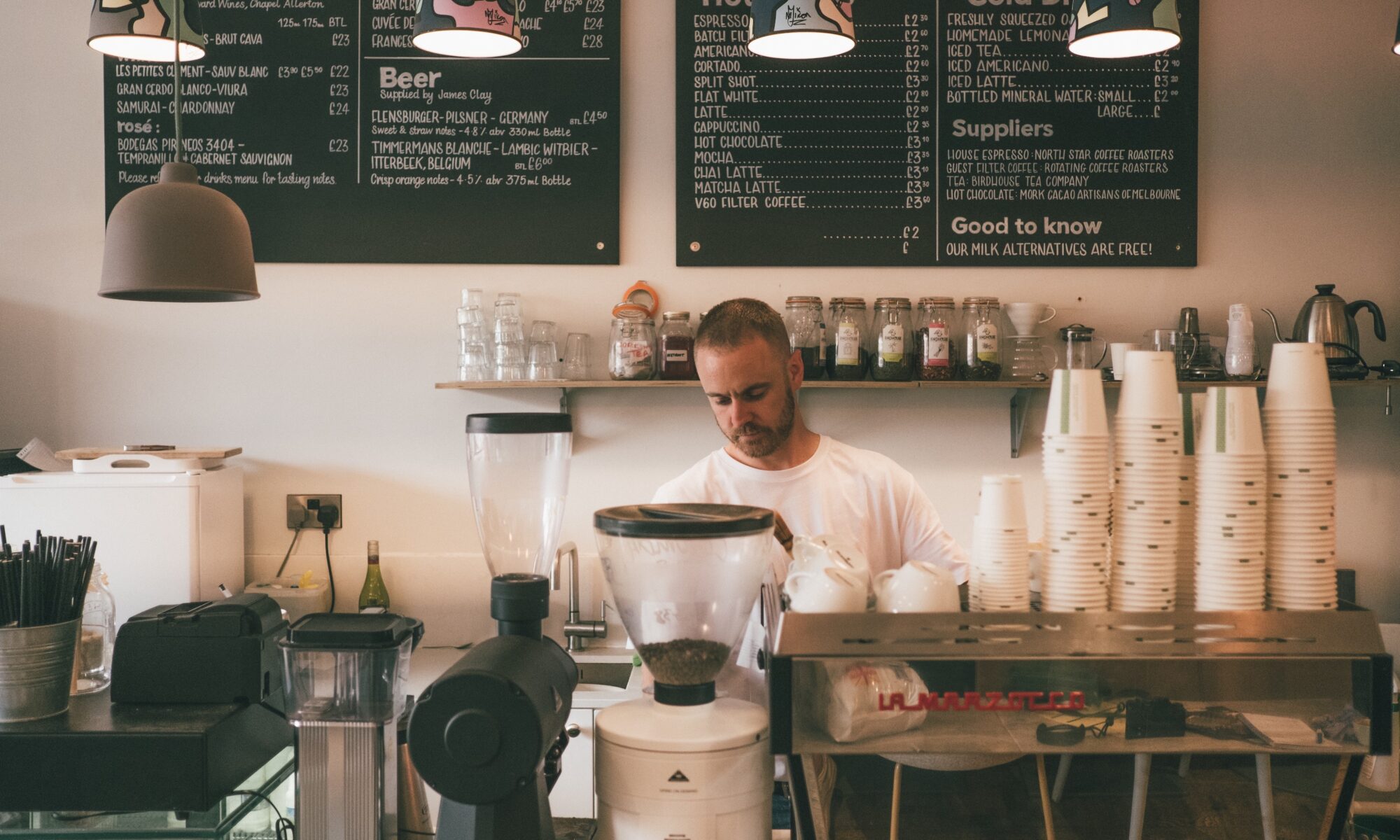 Man stands behind a large espresso machine inside a cafe surrounded by coffee cups